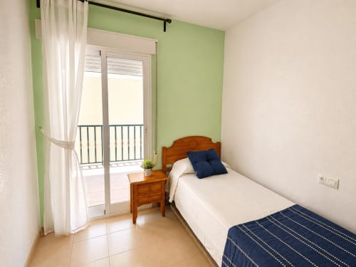 Dormitorio luminoso con pared de acento verde, cama de madera, almohadas azules y acceso a balcón en Apartamentos El Calón Playa