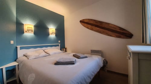 Modern bedroom with blue accent wall, white bed, nautical surfboard decor, and sconce lighting at Cap Coz apartment in La