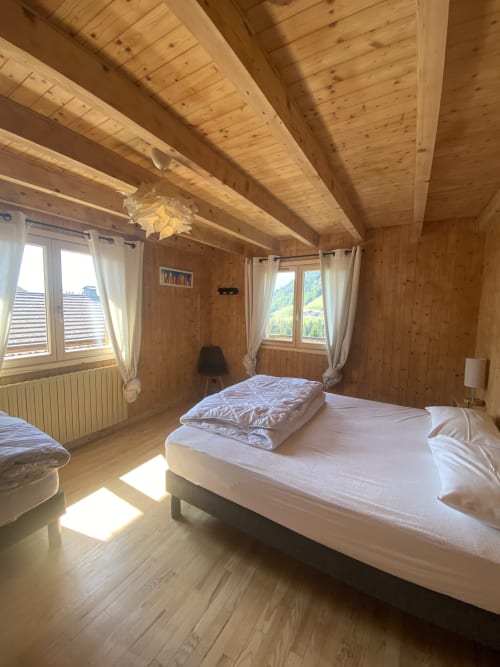 Bright wooden chalet bedroom with exposed beams, white bed, mountain views through windows, and natural lighting in