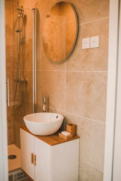 Salle de bain moderne du cottage Bobochic à ART DE VOYAGER, Etretat, avec miroir rond, vasque blanche, meuble en bois et carrelage