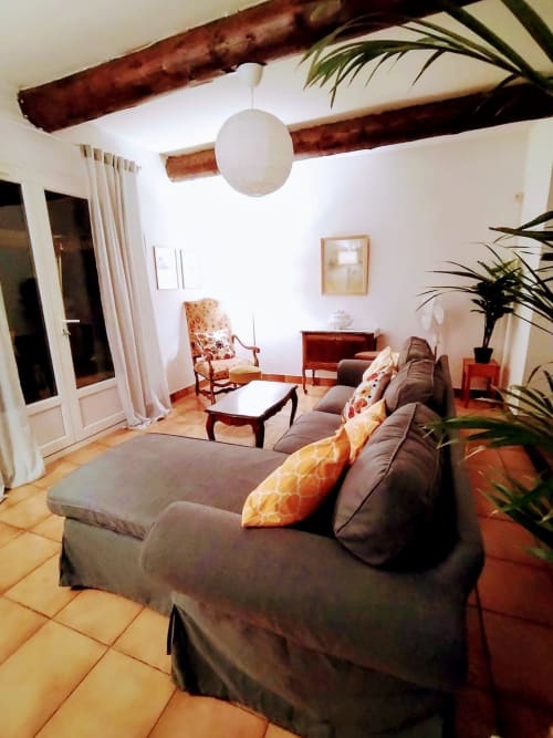 Spacious living room with exposed beams, gray sofa, wooden furniture, potted plants, and terracotta tiles in French villa