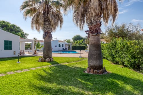 Jardín Las Golondrinas con palmeras, césped exuberante y piscina con vistas a los apartamentos Cap d'Artrutx