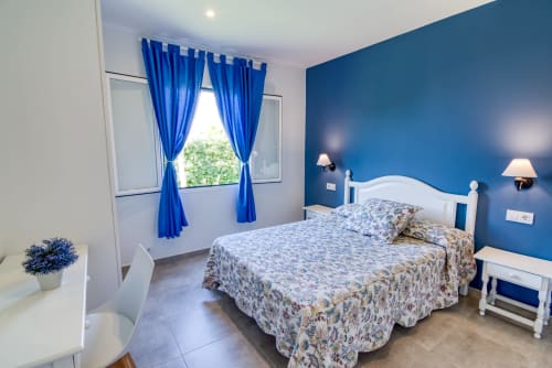 Dormitorio luminoso en Las Golondrinas con pared azul, cama blanca, cortinas azules y vistas al jardín en Cap d'Artrutx