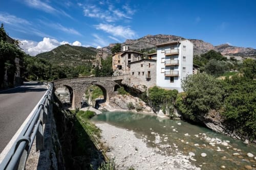 Puente de piedra histórico sobre río con edificios tradicionales y moderno edificio blanco en el valle de Montgat, Pirineos