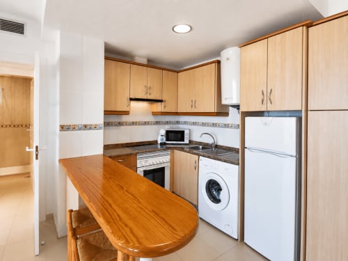 Cocina moderna con muebles de madera, electrodomésticos blancos, encimera de granito y mesa de comedor en Apartamentos El Calón Playa