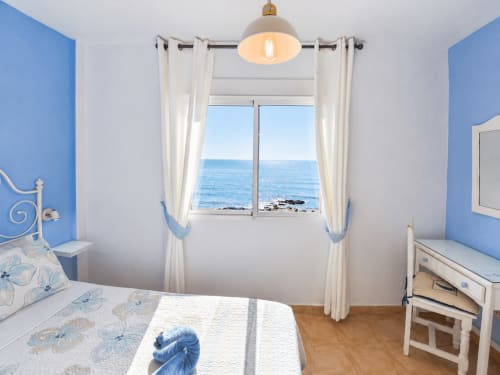 Bright bedroom with blue walls, sea view window, white curtains, and wooden floor at Apartamentos El Calón Playa