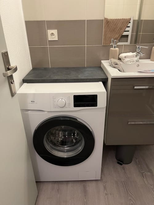 Salle de bains moderne avec machine à laver TCL blanche, plateforme de douche sombre, lavabo avec articles de toilette, et murs carrelés au Paillot de