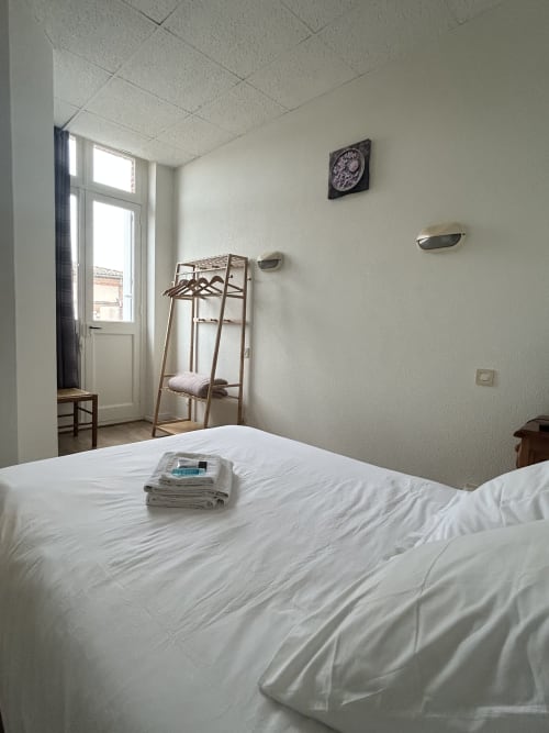 Chambre double propre à l'hôtel BDM de Montastruc-la-Conseillère avec literie blanche, portant vêtements, horloge murale et vue sur la ville