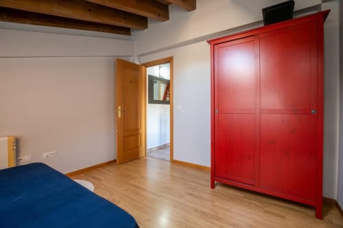 Luminoso dormitorio con vigas de madera, cama azul, puerta de madera y armario rojo llamativo en apartamento de Montgat