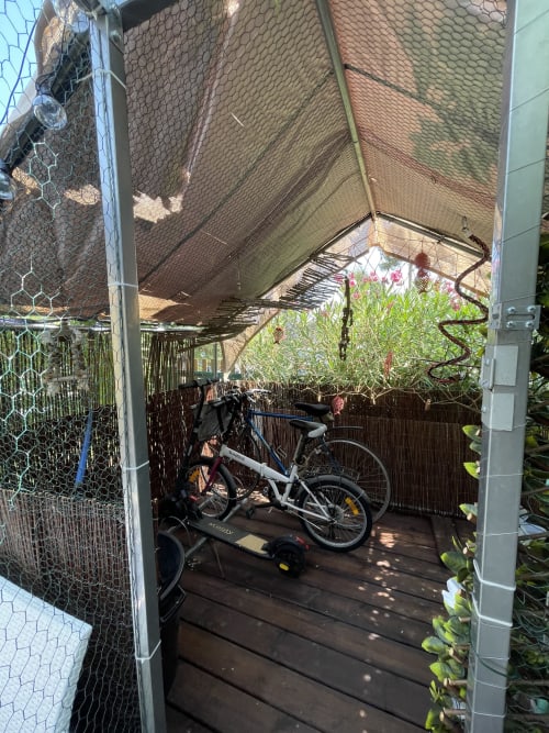 Abri à vélos couvert à l'appartement La Montagnette avec vélo blanc, plantes fleuries et terrasse en bois à Cap d'Agde