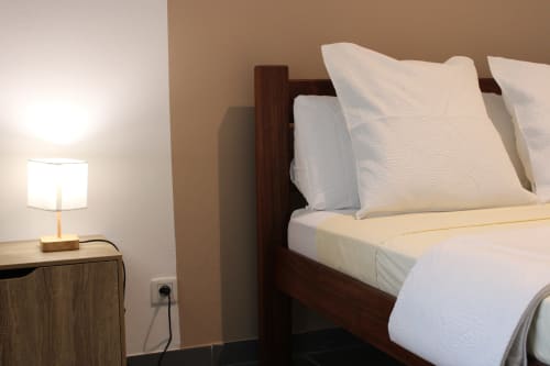 Chambre moderne aux Maisons En Or à Cayenne avec lit en bois foncé, oreillers blancs, lampe de chevet et murs crème
