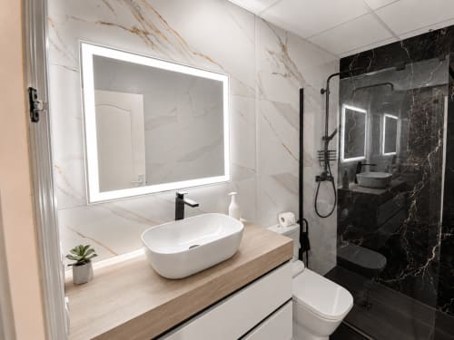 Baño moderno en Apartamentos El Calón Playa con lavabo de cerámica blanca, espejo iluminado, ducha de mármol negro y madera