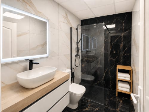 Baño moderno en Apartamentos El Calón Playa con lavabo blanco, mamparada de ducha en mármol negro y toallero de madera