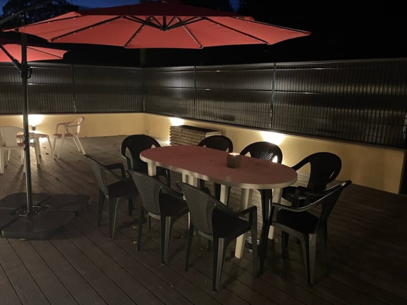Terrasse extérieure des Gites De Flaubert la nuit avec table à manger, chaises noires, parasol rouge et clôture métallique