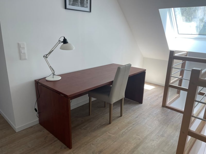 Lumineux espace de travail mansardé aux Gites De Flaubert avec bureau en bois foncé, lampe de bureau et fenêtre de toit vue sur Le Heron