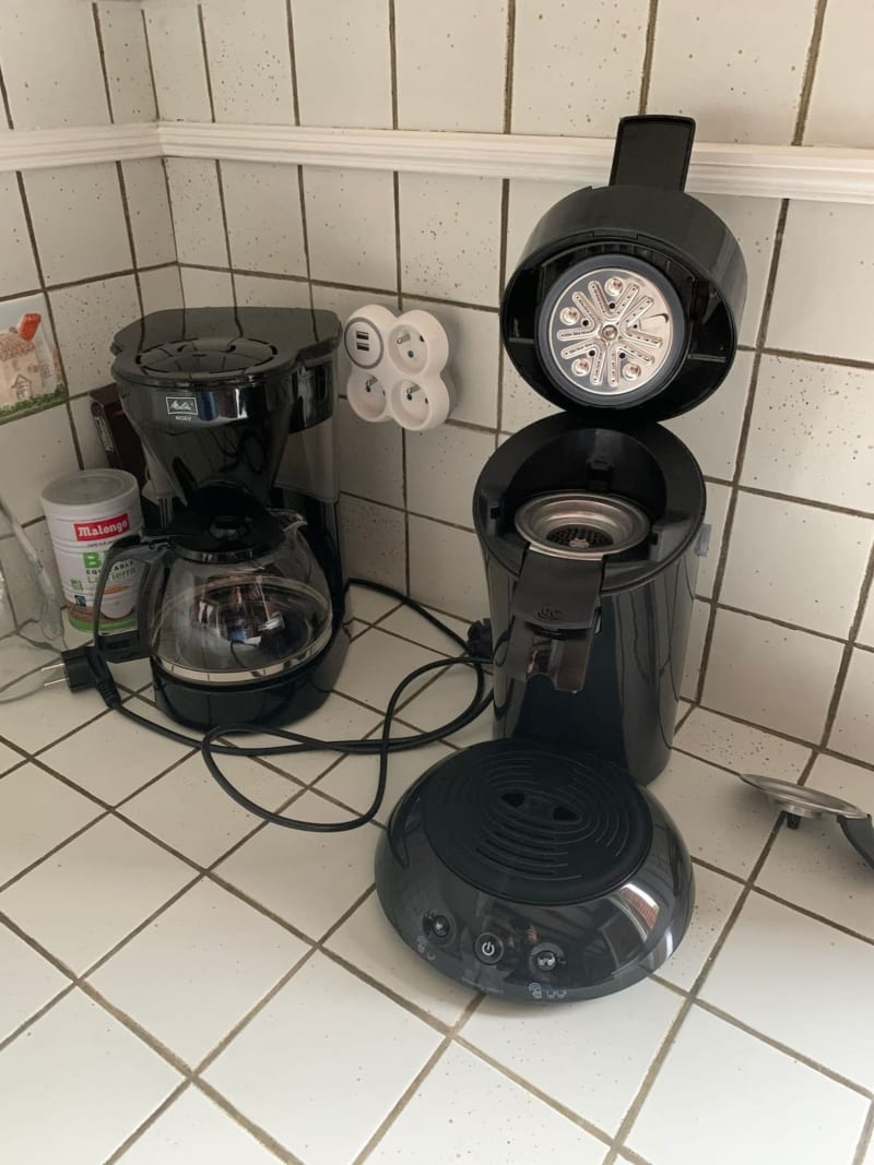 Comptoir de cuisine aux Gites De Flaubert avec cafetières noires, prises électriques et produits de nettoyage sur carrelage blanc