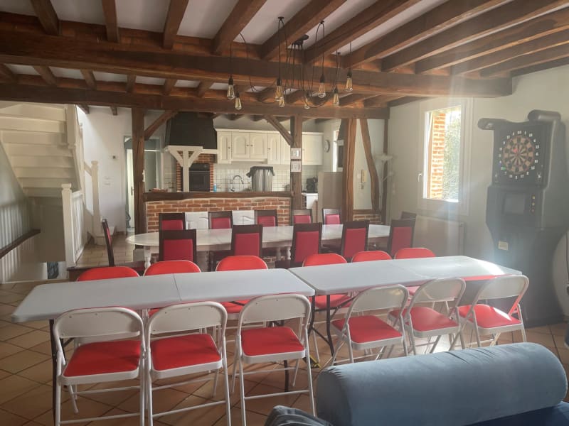 Spacieuse salle d'événements aux Gîtes De Flaubert avec poutres apparentes, chaises rouges, tables blanches et cuisine en briques.