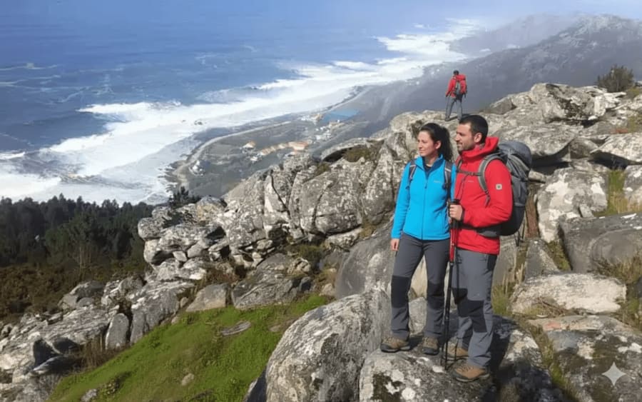 Senderistas con mochilas y bastones sobre rocas graníticas de la Serra da Groba con vistas al océano