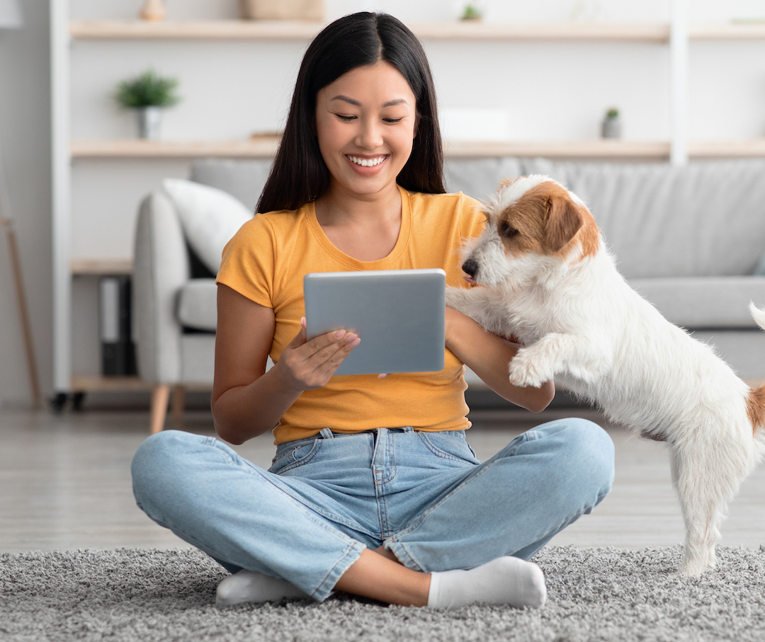 A woman in a yellow shirt sits cross-legged on the floor with an electronic device, playing with a puppy.