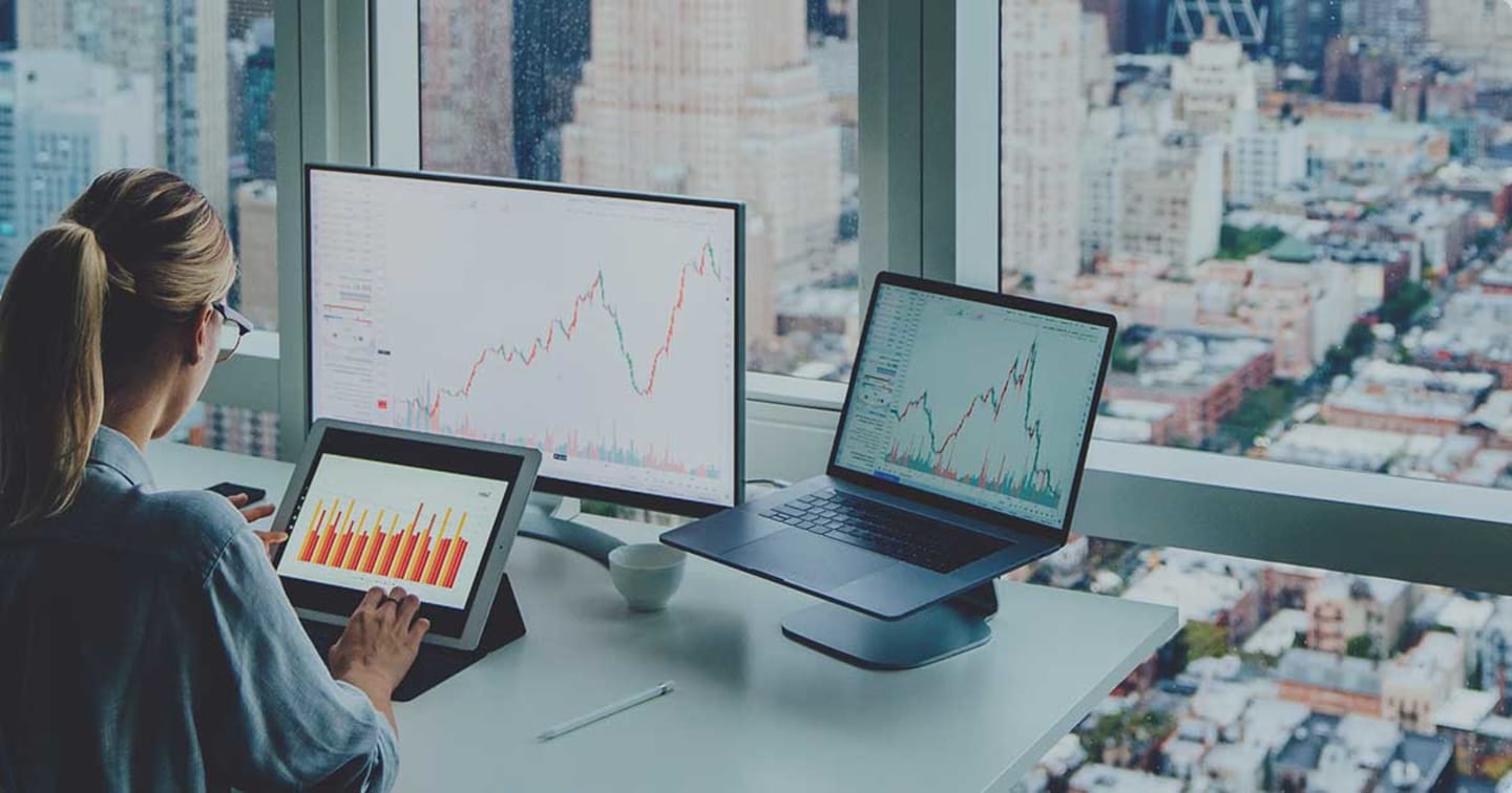 Women looking into tablet with a graph displayed and surrounded by a computer monitor and a laptop with graphs displayed as well.