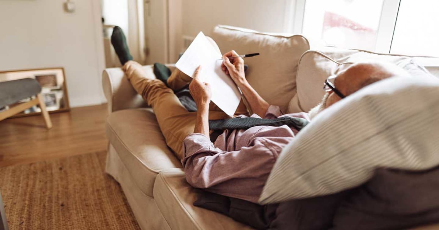 A man laying on his couch and writing on a piece of paper.