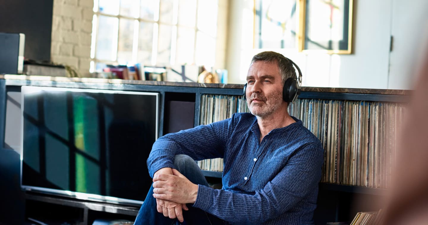 An older man listening to music on his headphones while leaning his back on shelves that hold his record collection.
