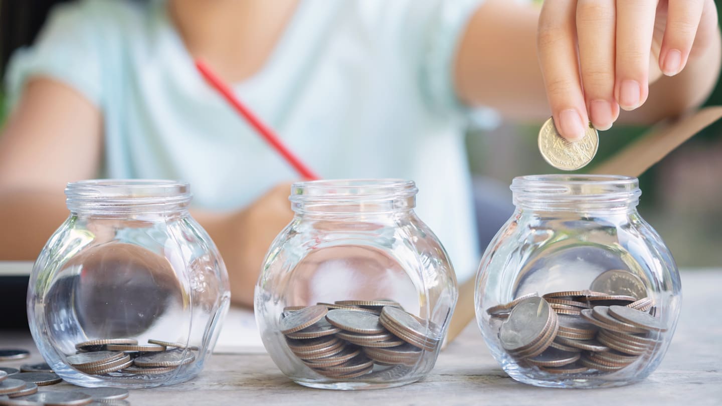 Three glass jars of the same size are in the forefront of the image, one with a small number of coins, one with a medium amount of coins and one with a larger amount of coins. A person’s hand is adding more coins to the jar with the most.