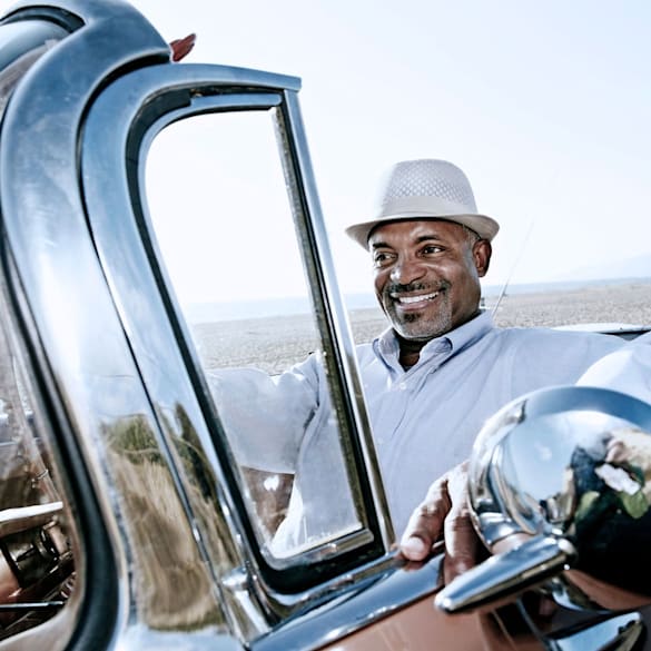 Man smiling while driving an older model convertible on a wide-open road.