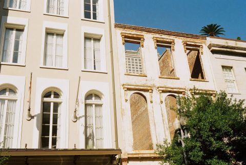 A row of older buildings, connected by just the remaining facade, its windows are a portal to the new development that can be seen in the background.