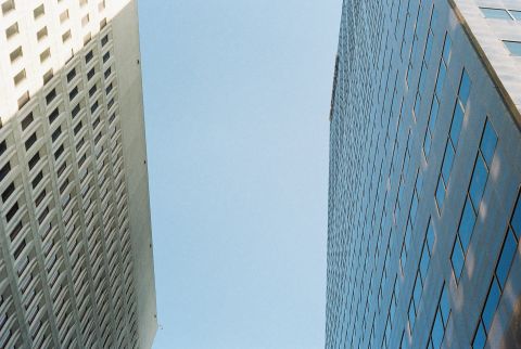 A path of blue sky opens between two high rises with rectangular glass windows.