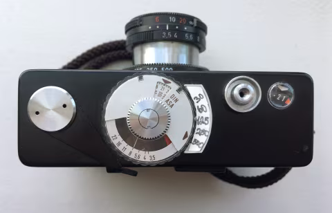 Top view of a Rollei 35B with the lens fully extended. The light meter is set up for an ISO/ASA 100 film via the small metal wheel at the top of the camera/middle of this image. The plastic transparent dial under it is rotated towards the 1/500th shutter speed, which I drew by hand with a pen on a small piece of thin cardboard. The needle shows that the safe aperture to use in these lighting conditions is 𝒇8, which I would have to change from 𝒇3.5, as marked on the lens barrel. You can almost see the shutter speed set to 1/500th as well, partially obstructed by the camera’s tiny body. At the tip of the lens, the distance is set to just under 10’, and on the left, you can see a counter, pointing to the 21st frame elapsed.
