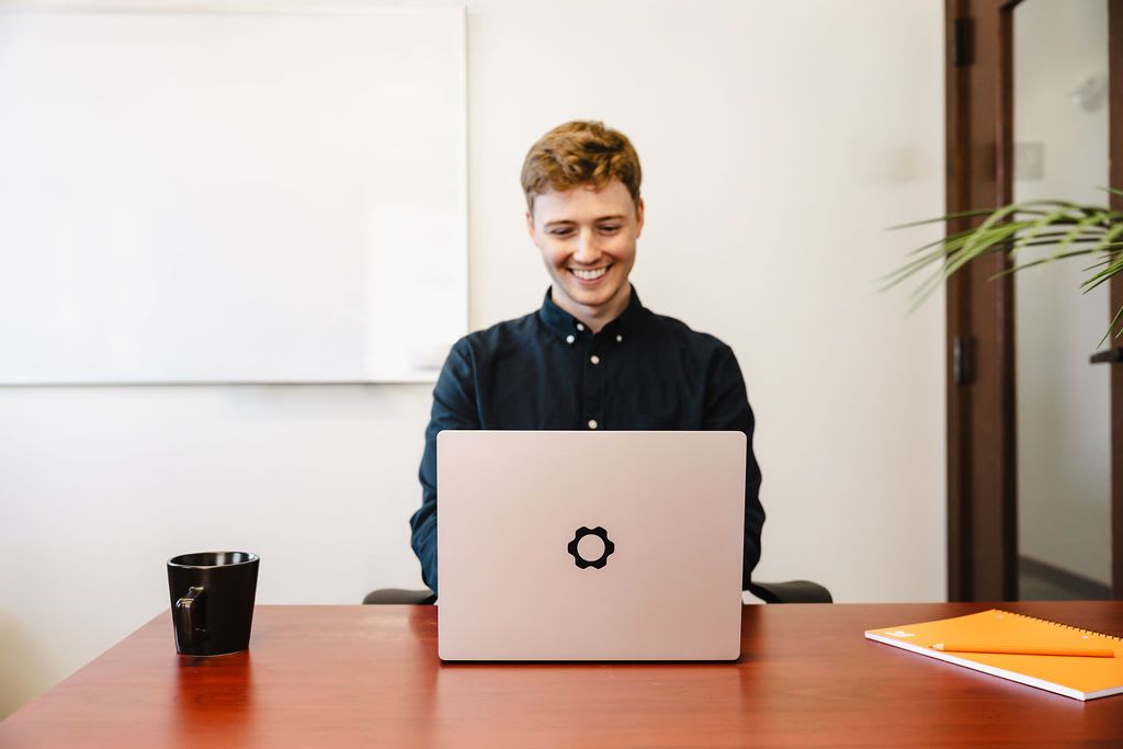 A netsuite consultant smiles while seated at a desk, working on a laptop.