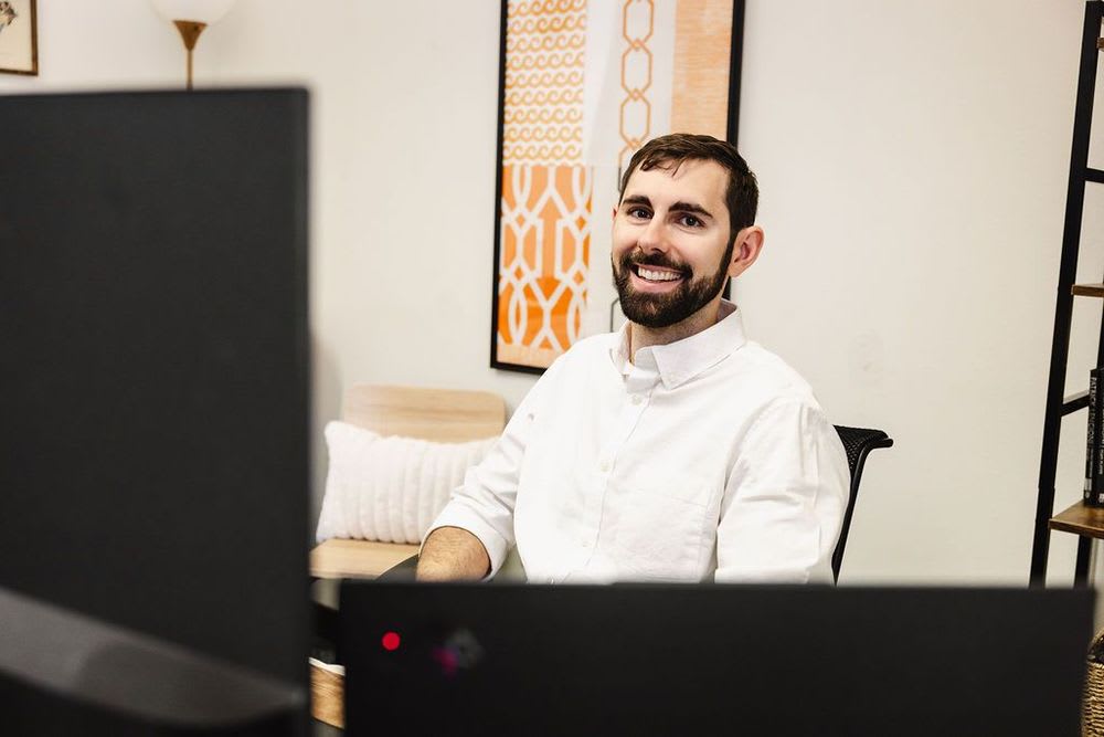 A smiling man with a beard sits at a desk, looking past his computer at the viewer.