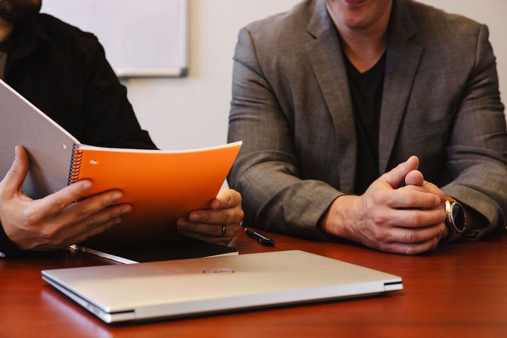 Two men seated at a table, reviewing a notebook, with a closed laptop and papers laid out in front of them.