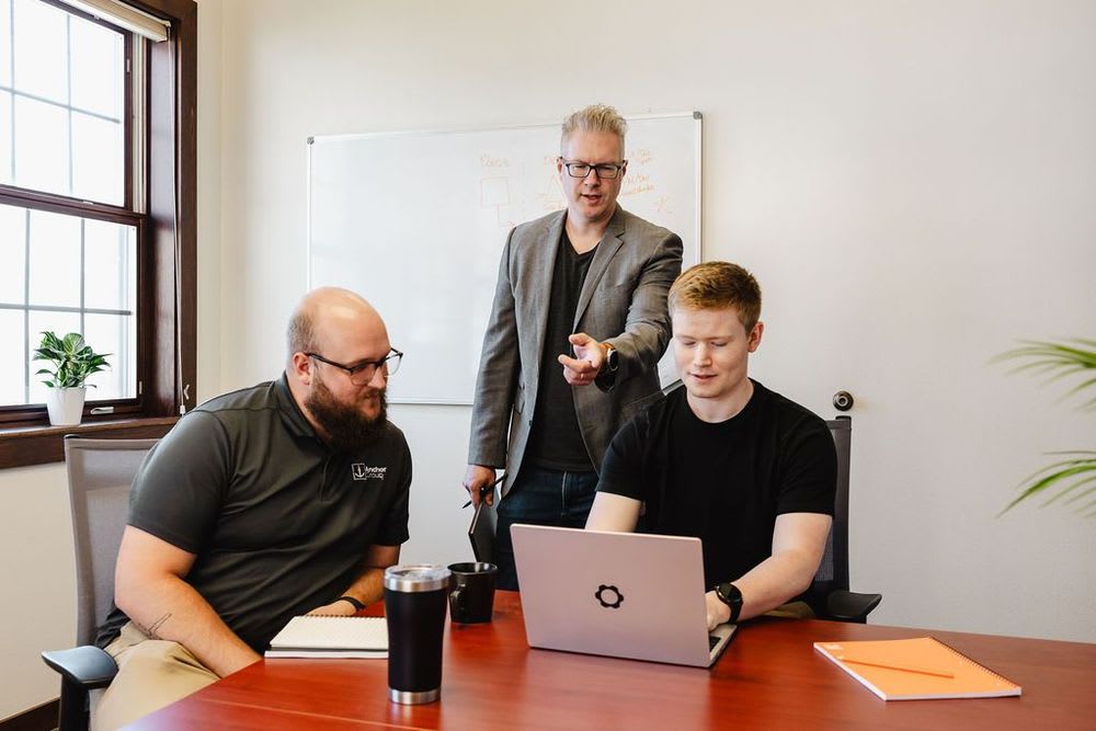 A group of three men gathered around a table, focused on a laptop during a discussion.