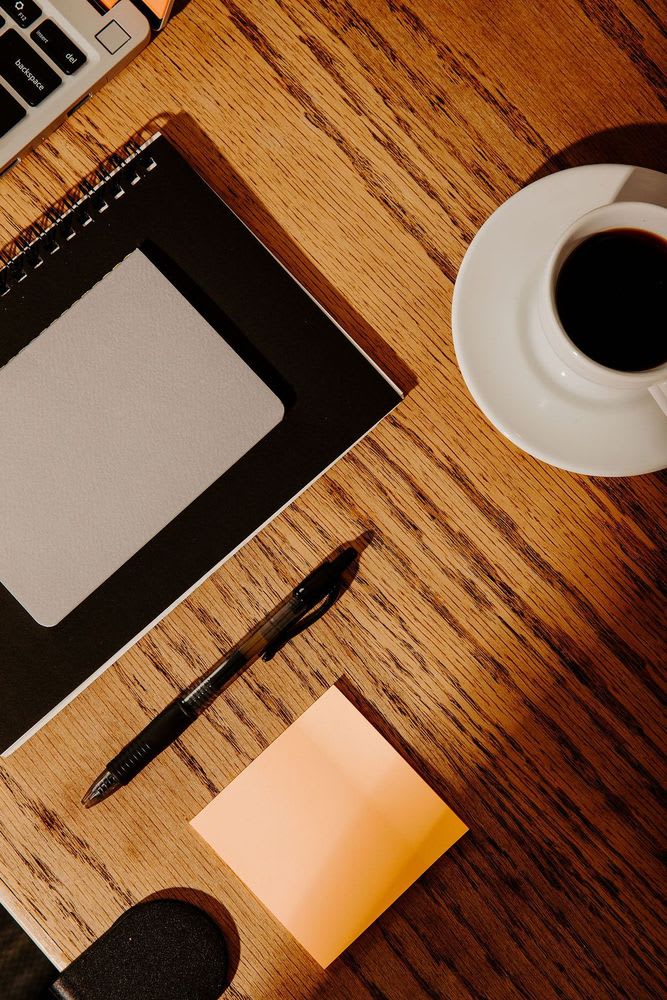 A laptop, coffee cup, pen, notepad, and phone arranged on a desk, suggesting a workspace setup for productivity.