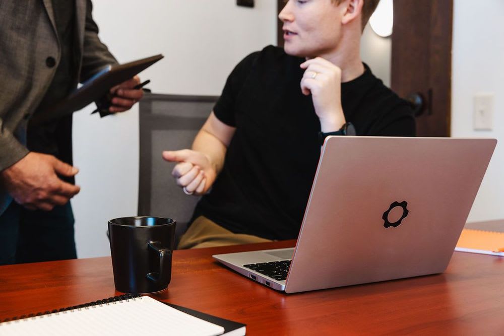 A person sitting at a desk with a laptop, speaking to another person beside him.