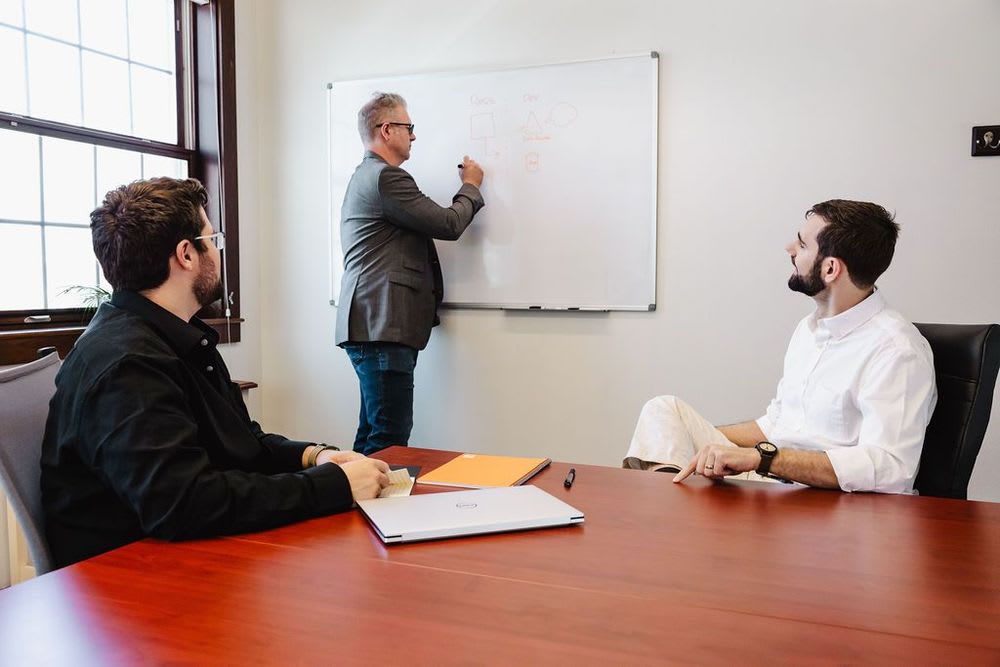 A person standing at a whiteboard while two men watch attentively.