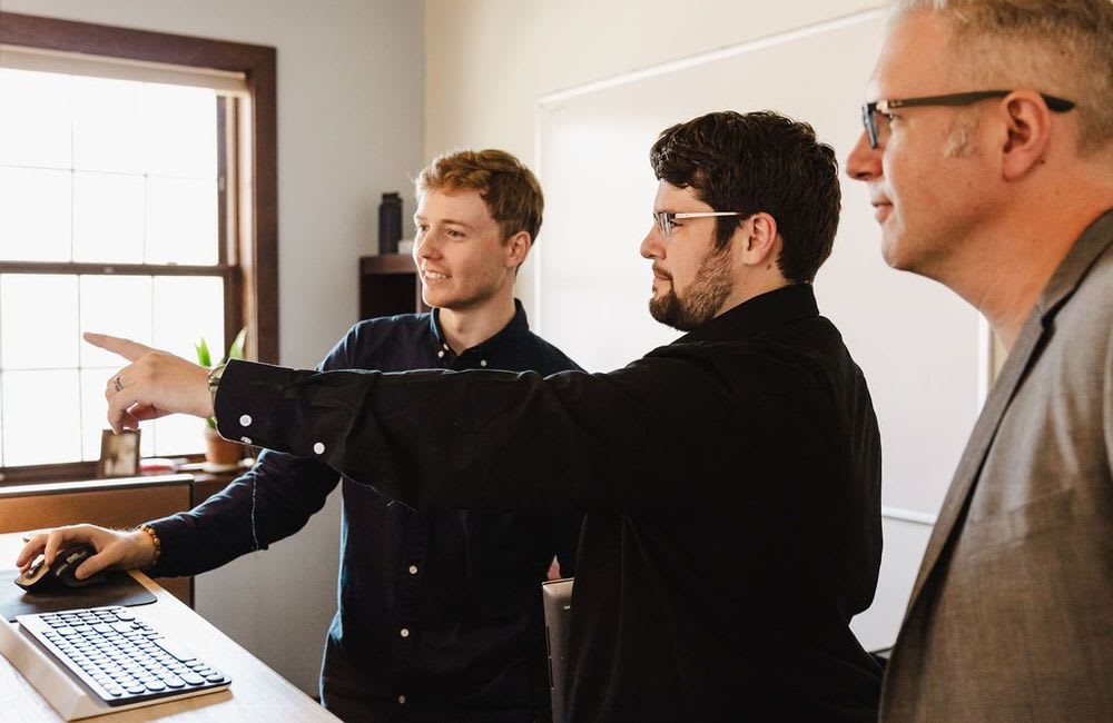 A person working at a computer while another watches, and a third points out specific points while smiling, illustrating collaboration and communication.