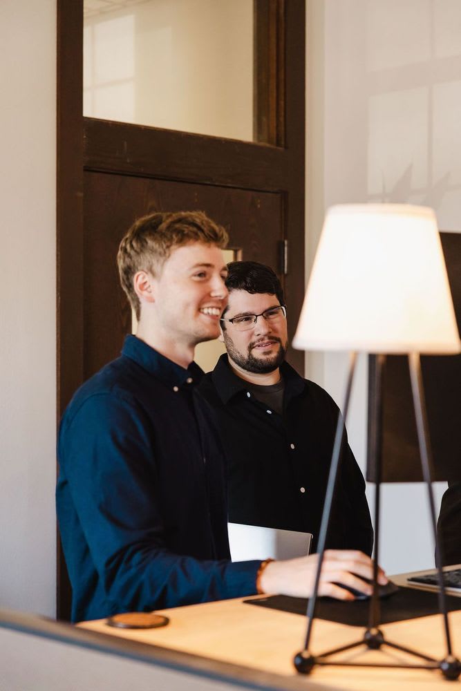 Two men are positioned by a desk featuring a lamp, discussing something intently.