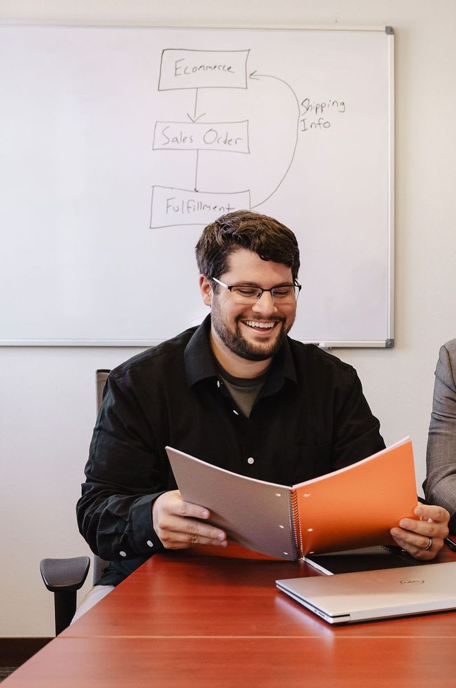 Two men seated at a table, discussing ideas with a whiteboard filled with notes and diagrams in the background.