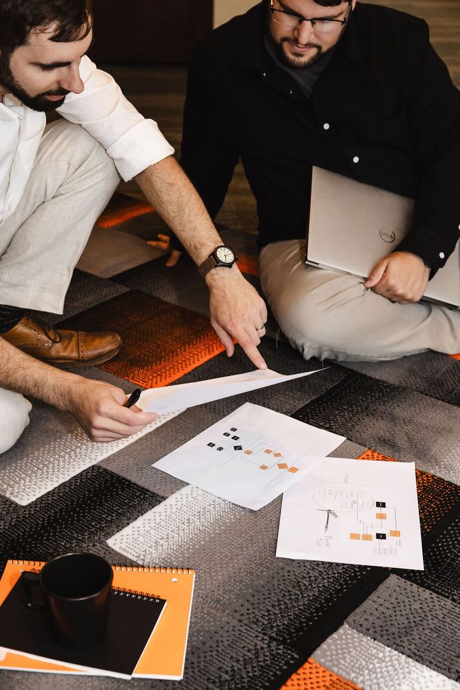 Two men seated on the floor, surrounded by papers and laptops, engaged in discussion or collaboration.