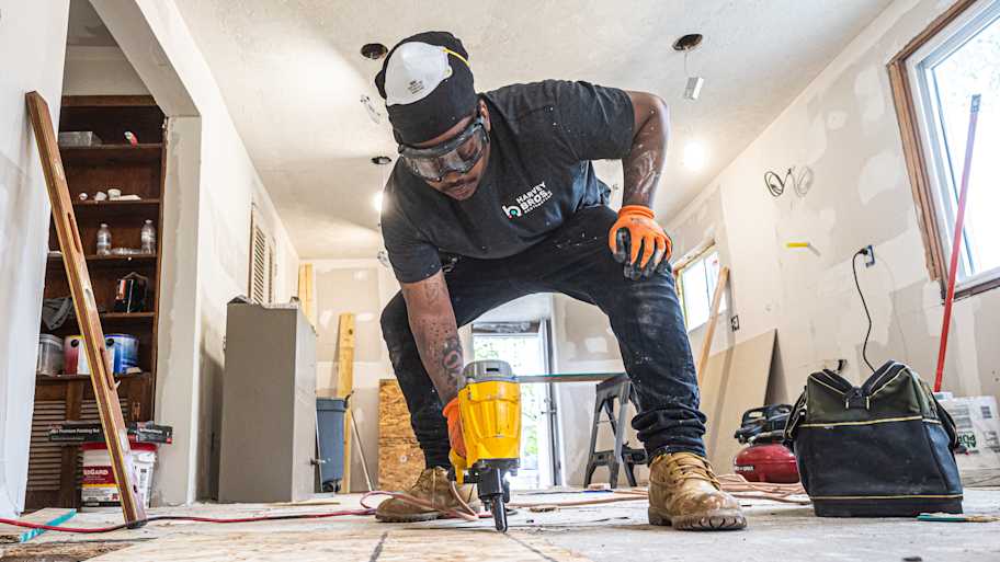 Construction worker using a nail gun to secure flooring during a home renovation, wearing safety gear and gloves.
