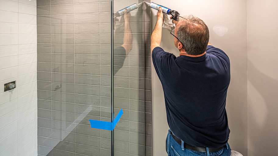 Contractor sealing the top edge of a glass shower door with caulk, ensuring a waterproof finish in a tiled bathroom.