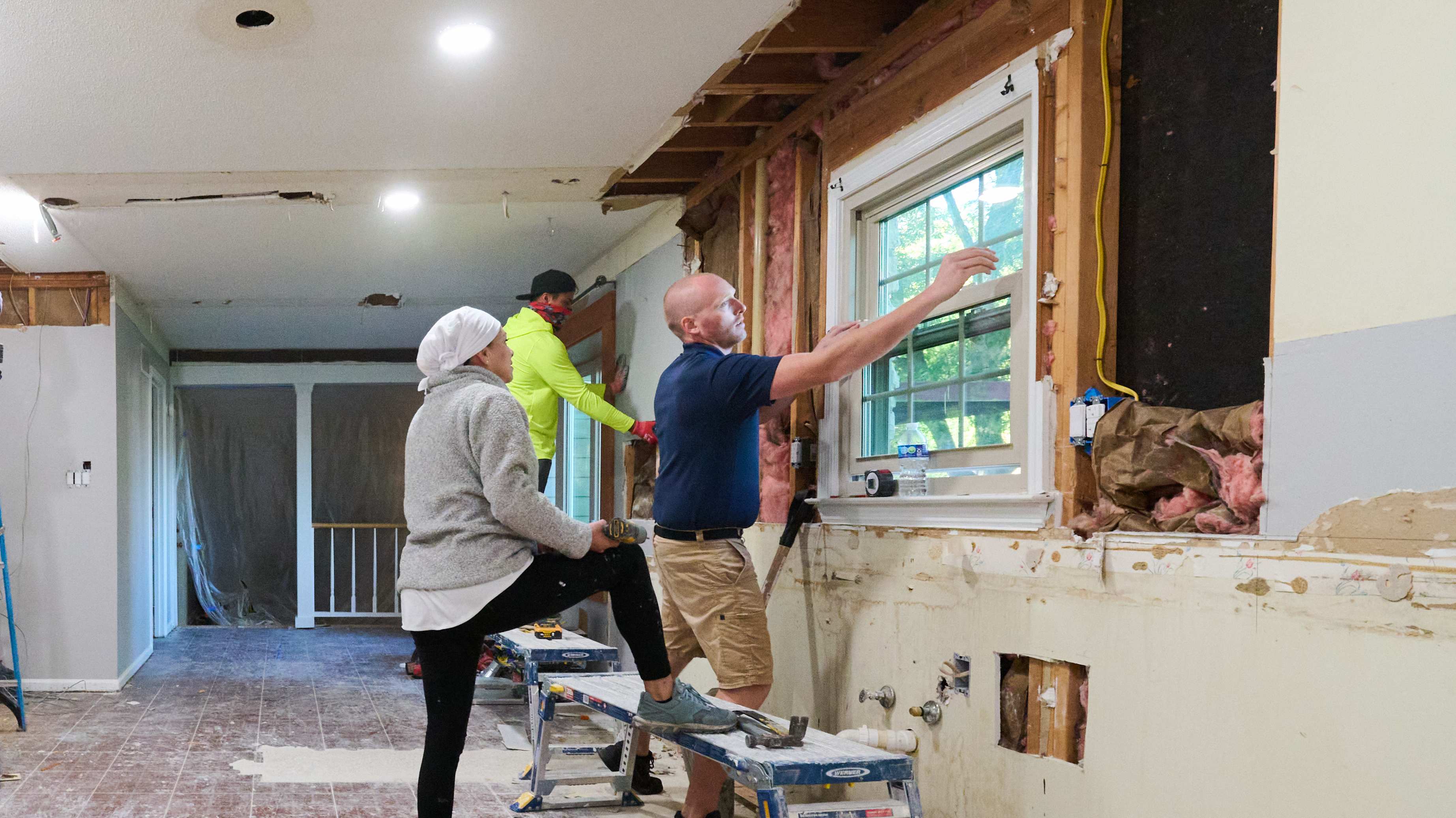 A renovation team works on walls and ceiling; one person is on a ladder while others remove drywall near a window and inspect progress.