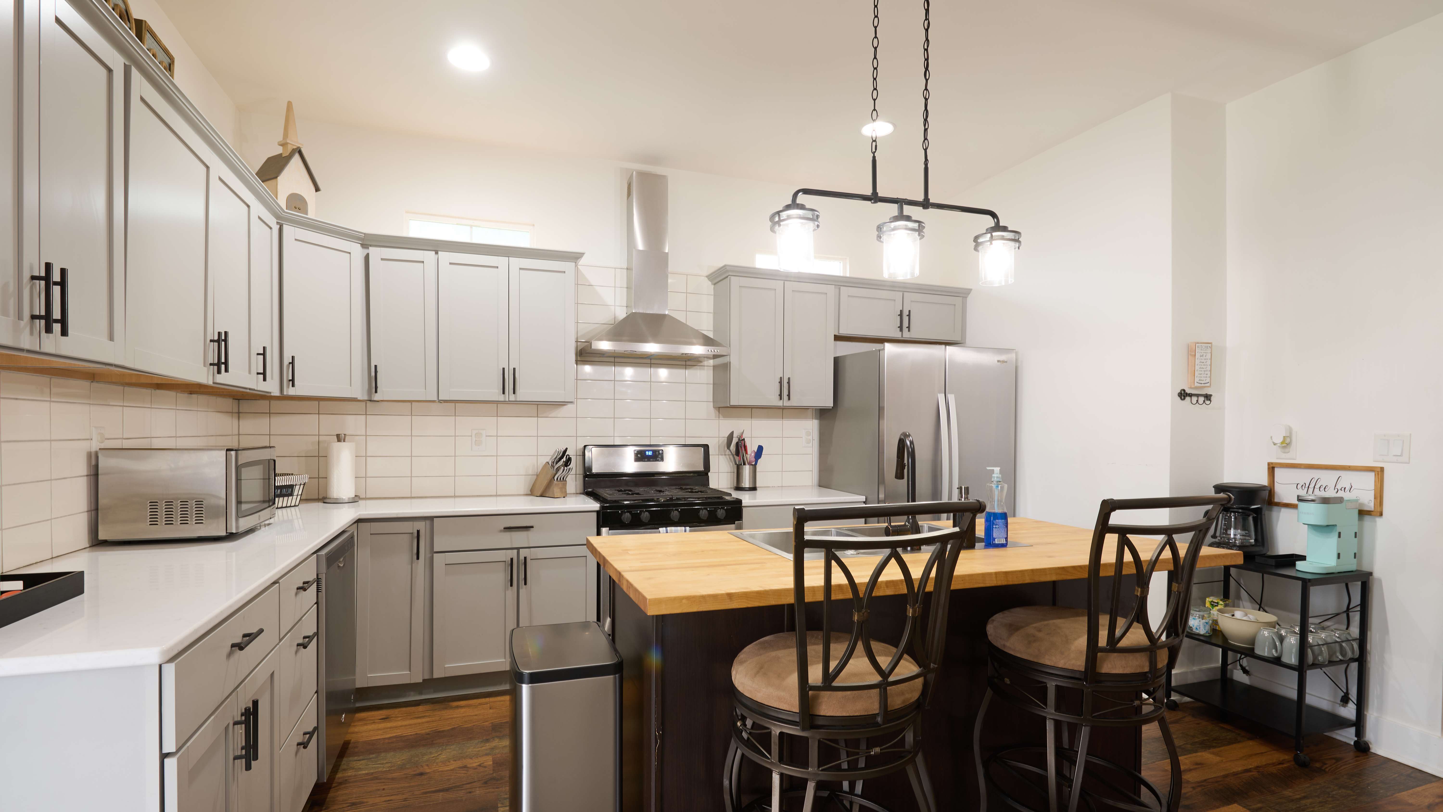 A modern kitchen with gray cabinets, a center island with wooden countertop and bar stools, stainless steel appliances, and pendant lighting.