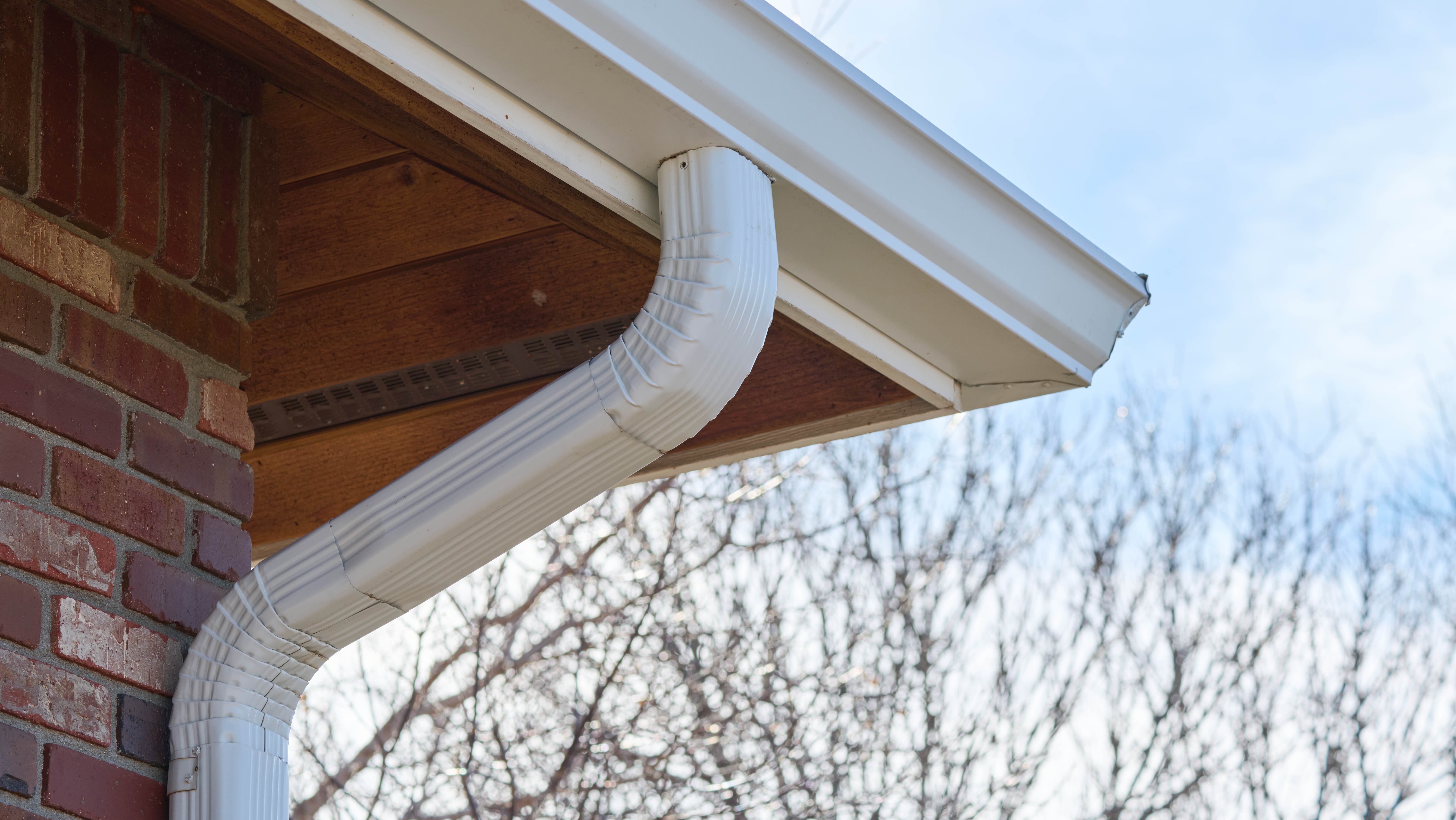 White downspout connected to seamless gutter system on brick home, directing rainwater away from the roof structure.
