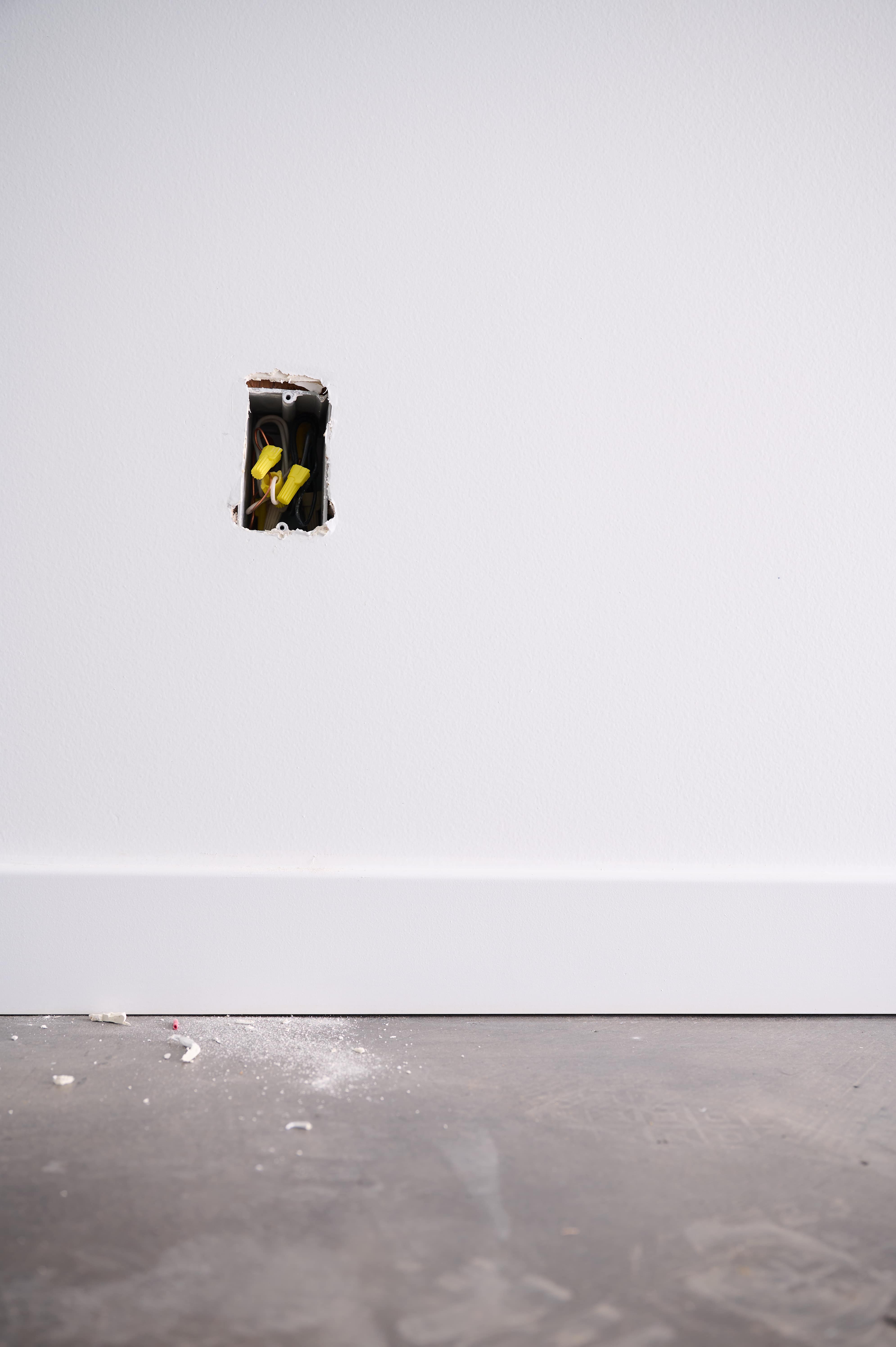 An open electrical outlet with exposed wires is set in a white wall, with drywall dust on the floor, showing ongoing electrical installation work.