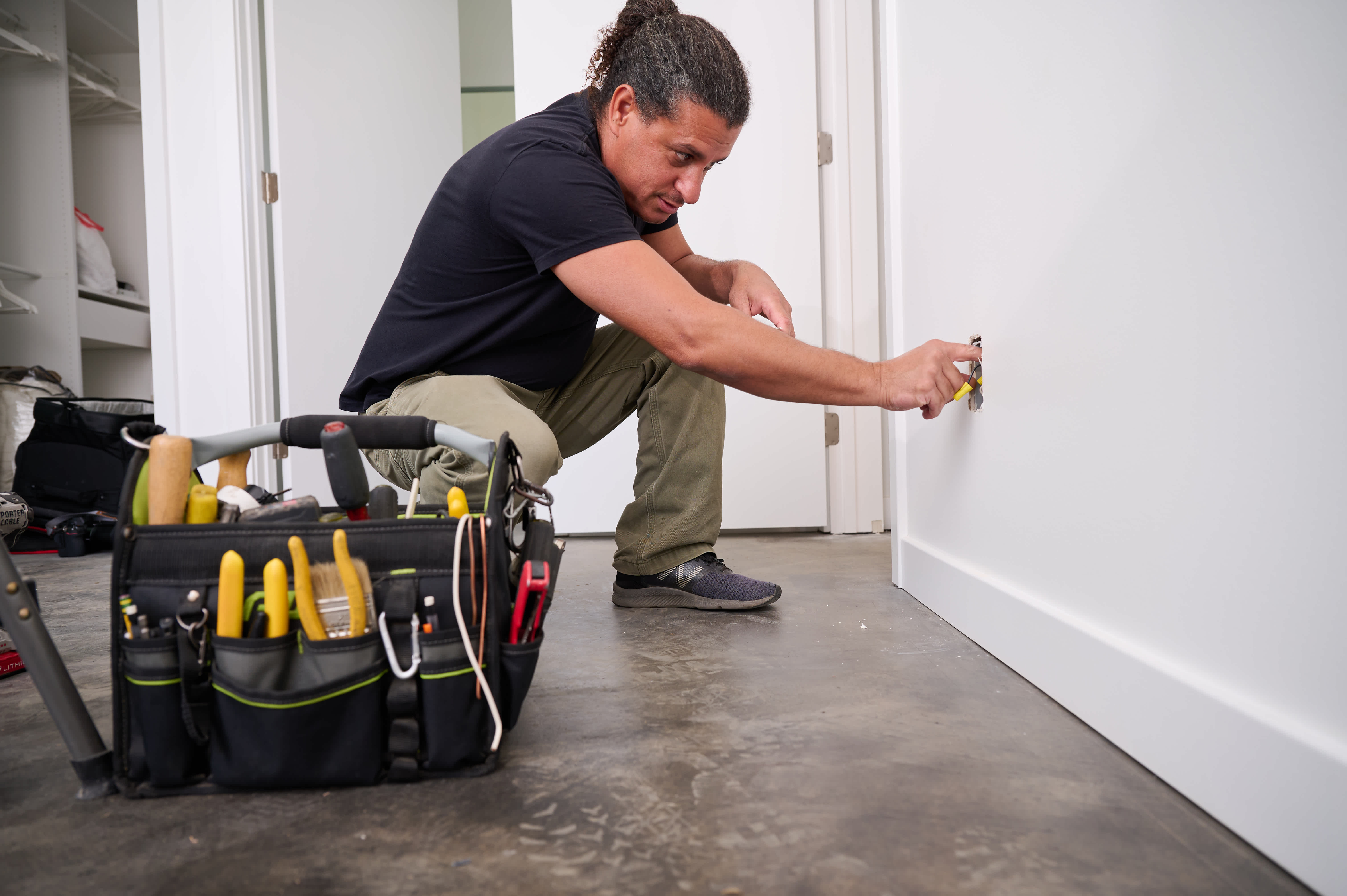 A technician kneels on the floor while working on an electrical outlet installation, with a tool bag full of equipment nearby for home repairs.
