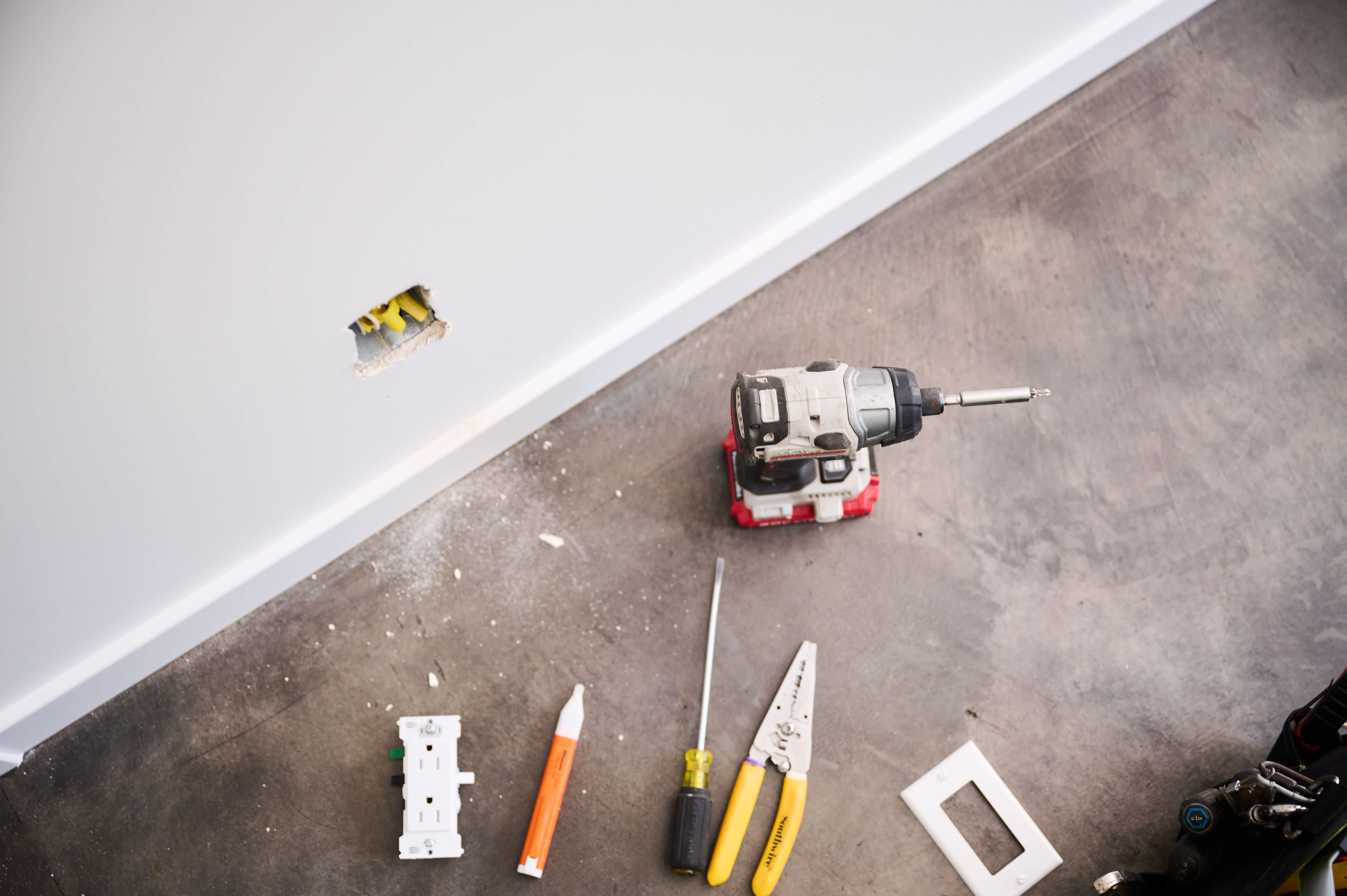A set of tools, including a power drill, screwdriver, wire stripper, and electrical socket, are neatly arranged on a concrete floor beside a wall opening.